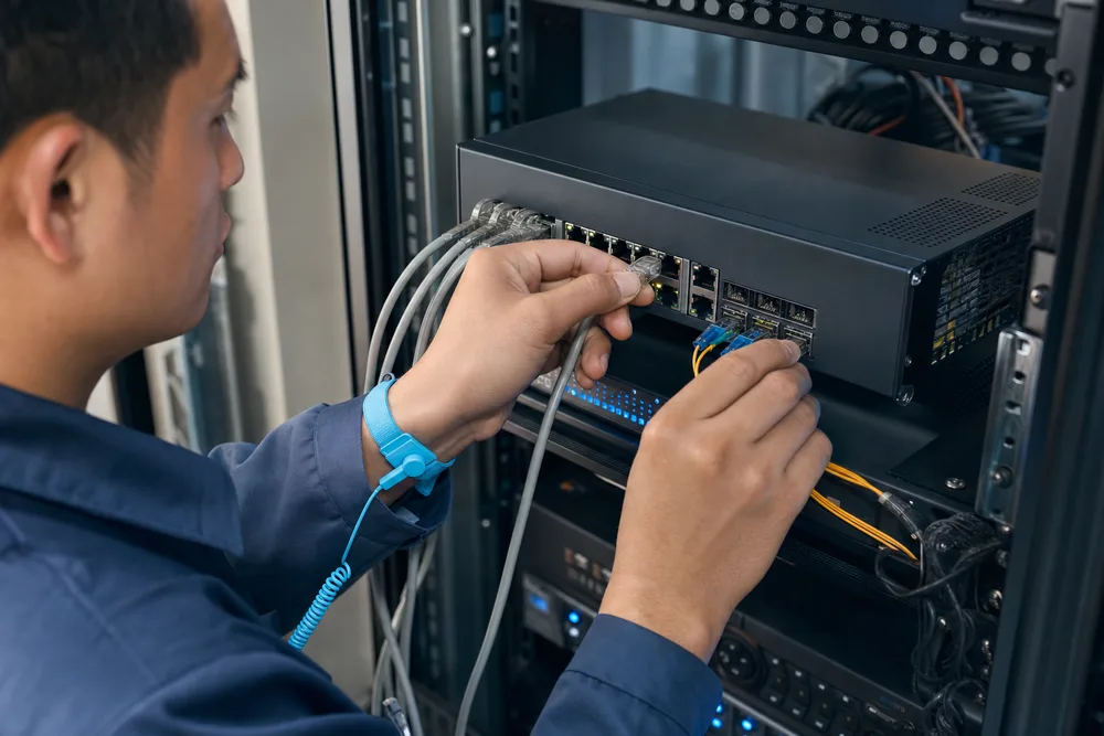 Network technician in a China CCTV equipment room checking copper RJ45 and fiber uplink connections on a compact Ethernet switch
